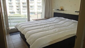 Une chambre d’hôtel avec un grand lit, une fenêtre donnant sur un immeuble et une table de chevet avec une lampe.