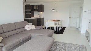 Een moderne woonkamer met een grijze modulaire bank, een klein wit tafeltje met stoelen, en een keuken in de achtergrond.