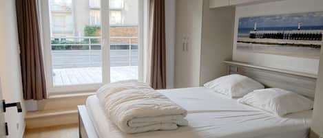 A hotel room with a large bed, a window with curtains, a painting above the bed, and a view of the outside balcony.