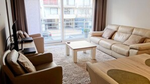 Een moderne woonkamer met een bank, fauteuil en salontafel. Er is een schuifdeur naar een buitenruimte met uitzicht op gebouwen en een geparkeerde auto.