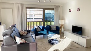 Un salon moderne comprenant un canapé, deux fauteuils, un téléviseur et un balcon offrant une vue sur les immeubles.