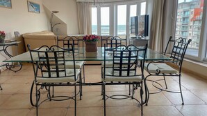 Een eetgedeelte met een glazen tafel en metalen stoelen, een bank, een televisie en uitzicht op zee door grote ramen.