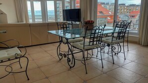Een moderne eethoek met een glazen tafel, metalen stoelen, een flatscreen televisie en een uitzicht op de skyline door grote ramen.