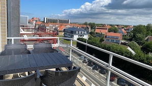 Un balcon doté de meubles en osier, donnant sur une rue animée par la circulation des voitures et bordée d’immeubles.
