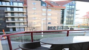 A balcony with wicker chairs and a glass table, overlooking a modern building with multiple balconies and a glass facade. - Apartment in Nieuwpoort near the Beach (Nieuwpoort)