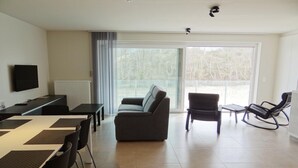 Una sala de estar moderna con un sofá, una mesa de comedor con sillas, un televisor montado en la pared y un amplio ventanal corredero que da a un área exterior.