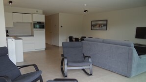 Una sala de estar moderna con un sofá gris, un comedor con una mesa y sillas, y una cocina con armarios y electrodomésticos blancos.