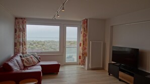 A living room with a sofa, a TV, and a view of the sea through large windows. - Apartment in Nieuwpoort by Sandy Beaches (Nieuwpoort)
