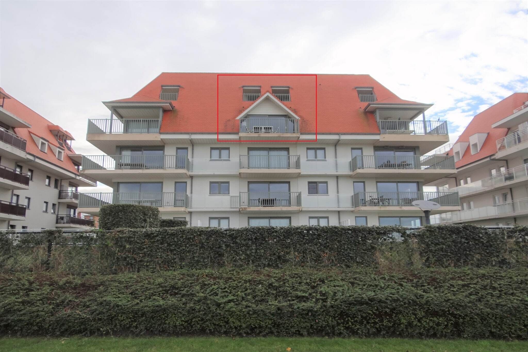 A multi-story residential building with balconies, a red roof, and a hedge in the foreground.