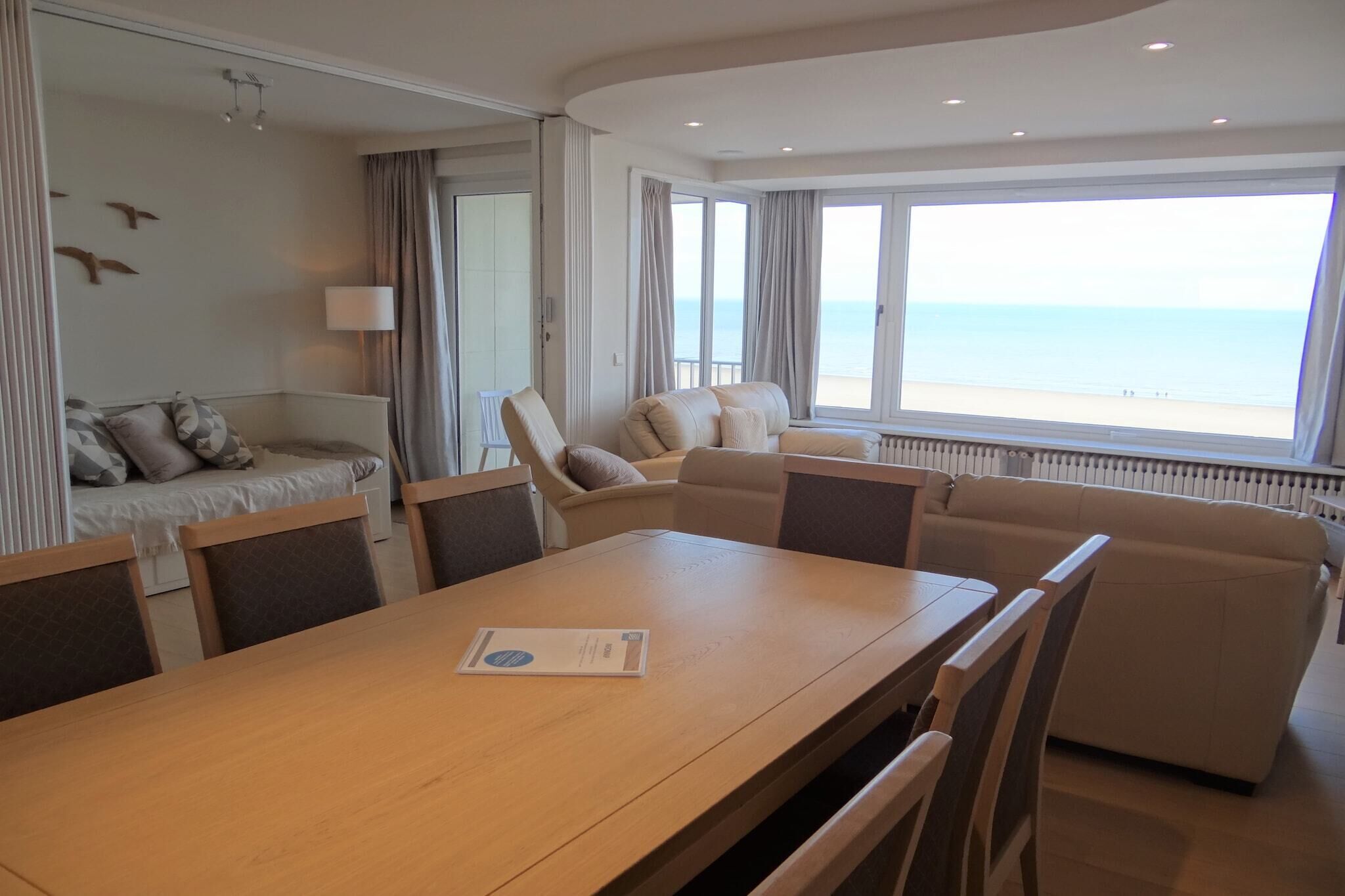 A room with a wooden dining table, beige sofas, and a view of the sea through large windows.
