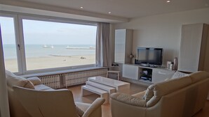Un salon moderne doté d’une grande fenêtre donnant sur une plage, d’un téléviseur à écran plat et d’un design minimaliste.