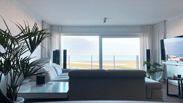 A modern living room with a large sofa, a coffee table, and a TV on a stand. There are two large windows with curtains, a plant, and a view of a distant landscape. - Apartment in Nieuwpoort for 4-people (Nieuwpoort)