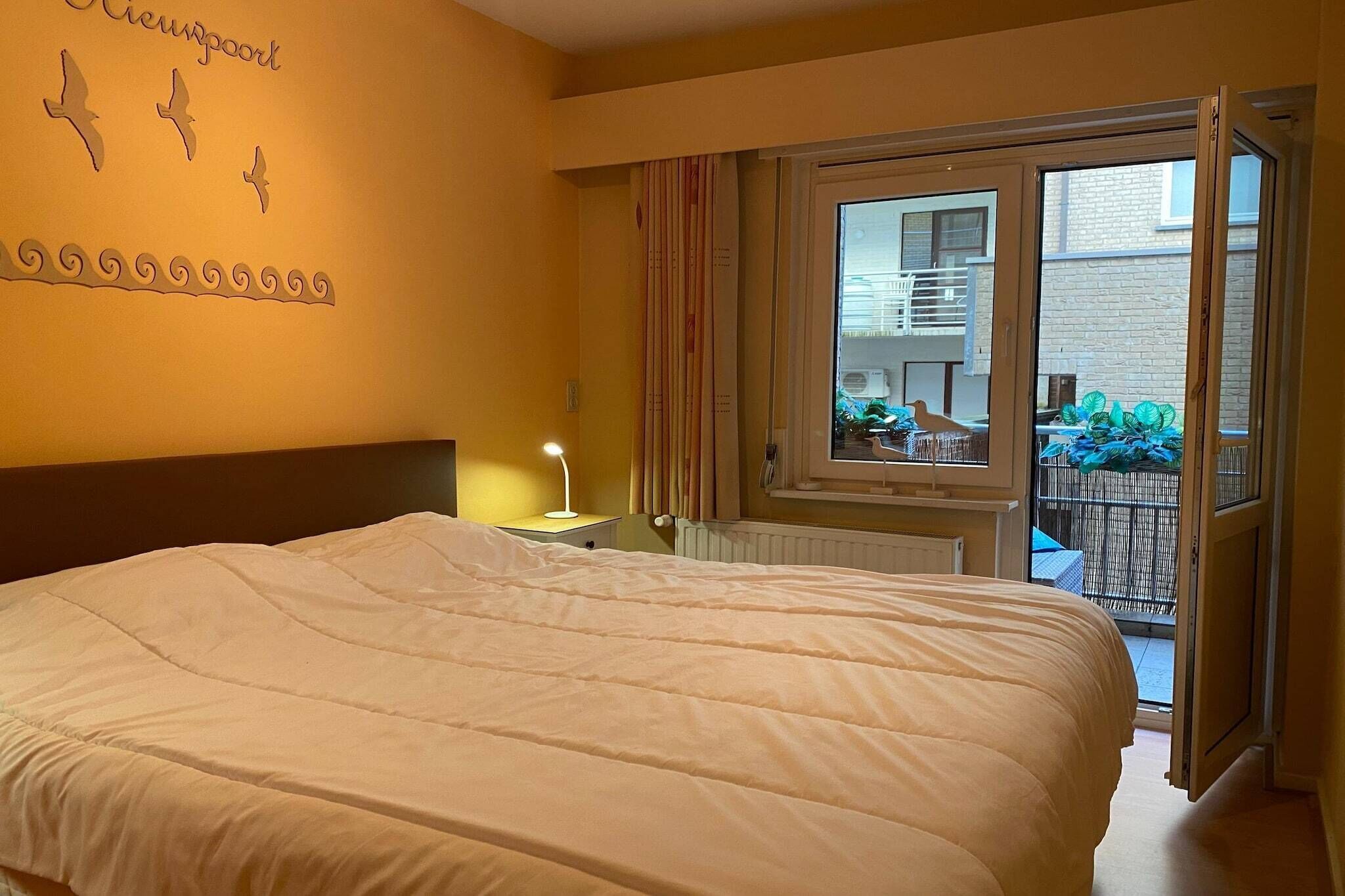 Ein Hotelzimmer mit einem großen Bett, einem Nachttisch mit Lampe, einem Fenster mit Blick auf den Balkon und einer Wand mit einem dekorativen Vogel- und Wirbelmuster.