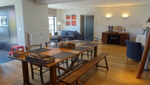 A spacious living area with a wooden dining table, benches, and a sofa. There is a kitchen area with a stove and a refrigerator. The room has a large window providing natural light. - Bright Attic with Sea Breeze (Nieuwpoort)