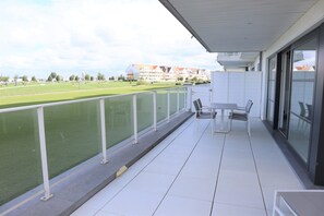 Un balcon donnant sur un terrain de sport, une table et des chaises, et une porte vitrée menant à une pièce.