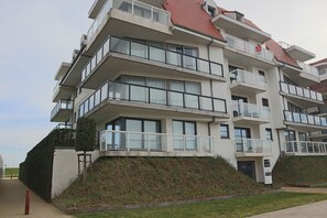 Un immeuble résidentiel à plusieurs étages, doté de balcons, d’un toit rouge et d’un espace verdoyant à l’avant.