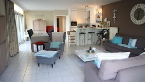 A modern living room with a grey sofa, a coffee table, and a dining area with a table and chairs. A kitchen with white cabinets and a large clock on the wall is visible in the background.