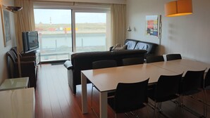 A modern hotel room with a large window, a sofa, a dining table, and a TV.