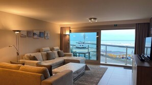 Een moderne woonkamer met een zithoek, een flatscreen televisie en uitzicht op zee door grote ramen.