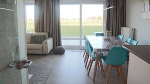 En moderne stue med sofa, spisebord med stole og udsigt til et grønt landskab gennem store vinduer.