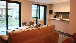 A modern living room with a sofa and two chairs, a kitchen with a sink and appliances, and large windows offering a view of greenery outside.