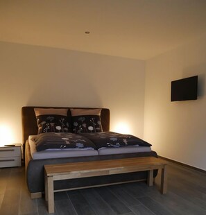 A bedroom with a bed, bedside tables, a bench, and a wall-mounted TV. - Appartement am See, 1-2 Pers (Haltern am See)