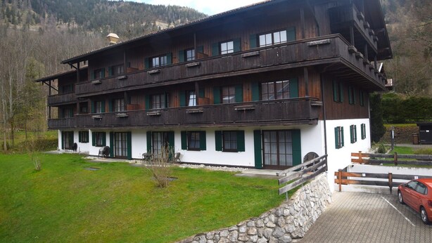 A two-story wooden building with balconies, a stone wall, and a parking area with a car. - Ferienwohnung in Bayrischzell (Bayrischzell)