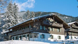 A multi-story wooden building with balconies, surrounded by snow-covered trees and mountains. - Ferienwohnung in Bayrischzell (Bayrischzell)