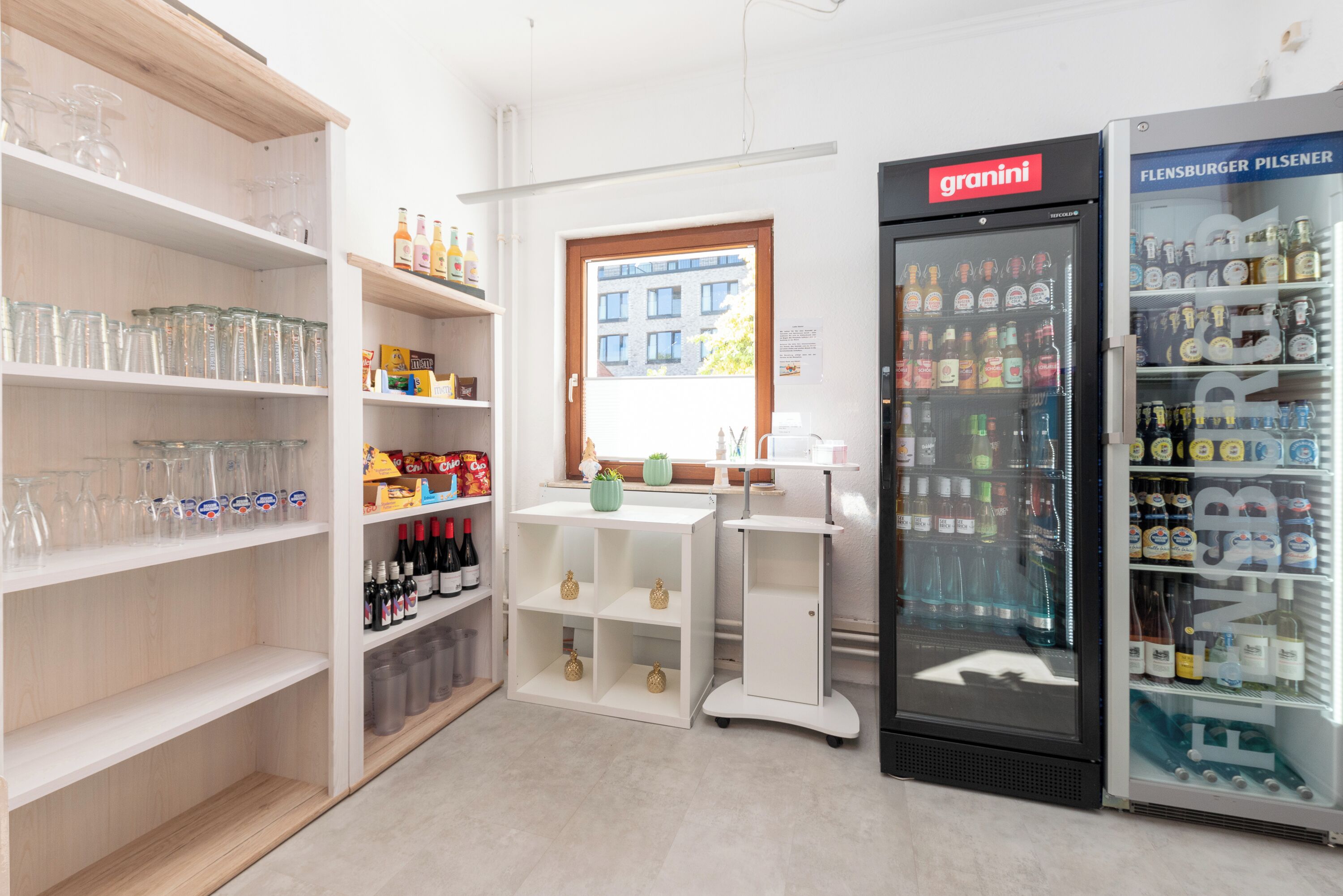 A room with shelves of glassware, a refrigerator stocked with beverages, and a window with a view of buildings.
