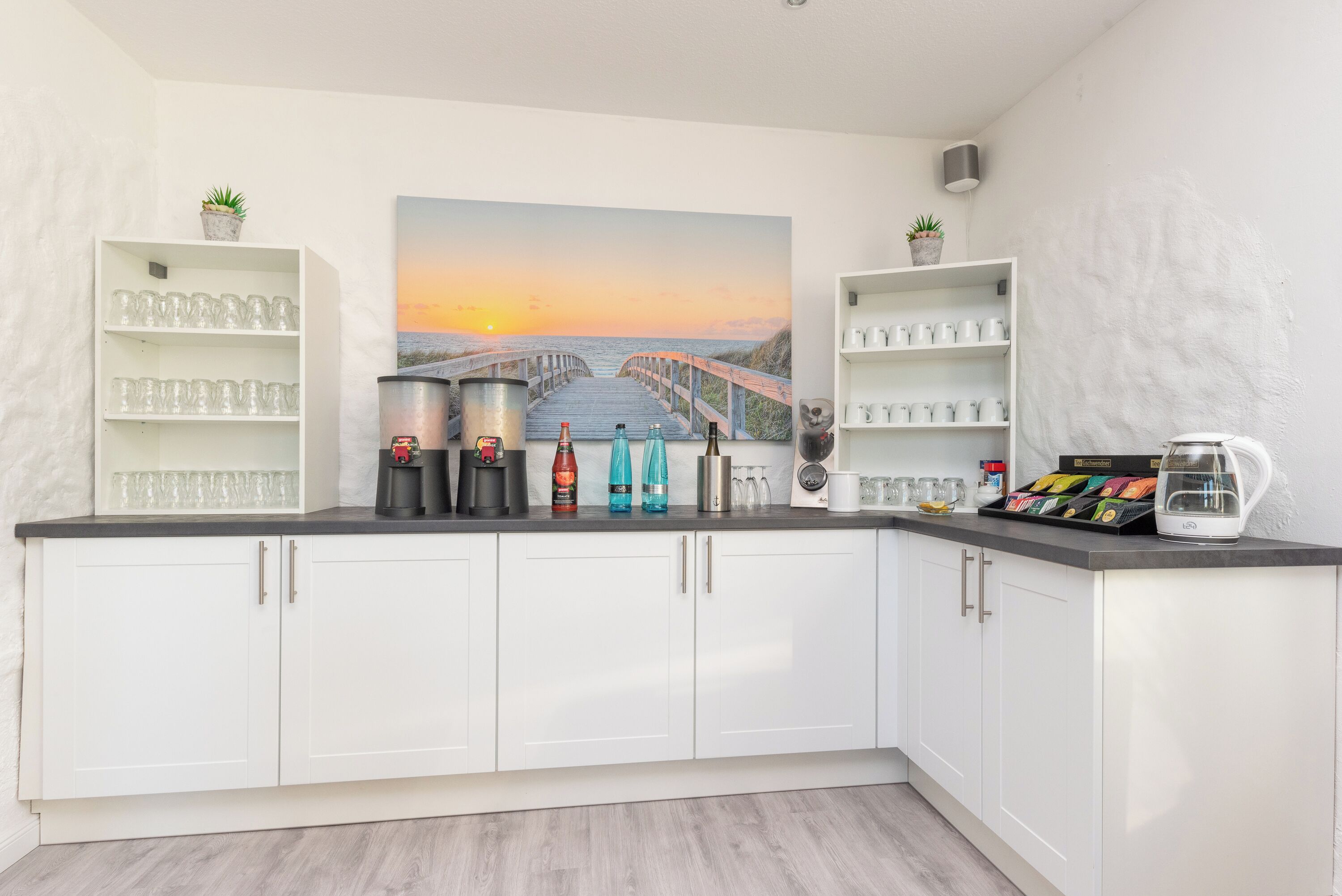 A modern kitchen with white cabinets, a coffee station, and a scenic sunset painting on the wall.