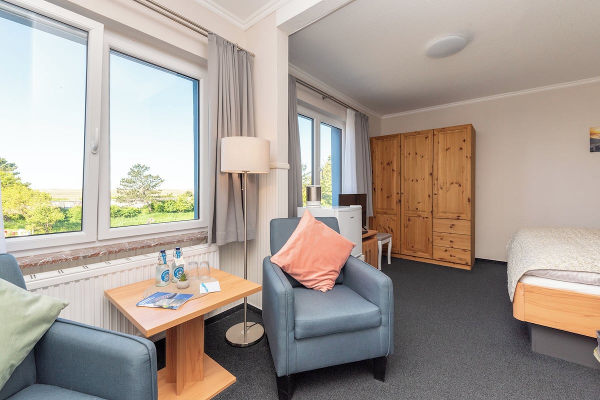 Une chambre d’hôtel avec un lit, un bureau avec une lampe, une chaise, une armoire et de grandes fenêtres donnant sur un beau paysage.