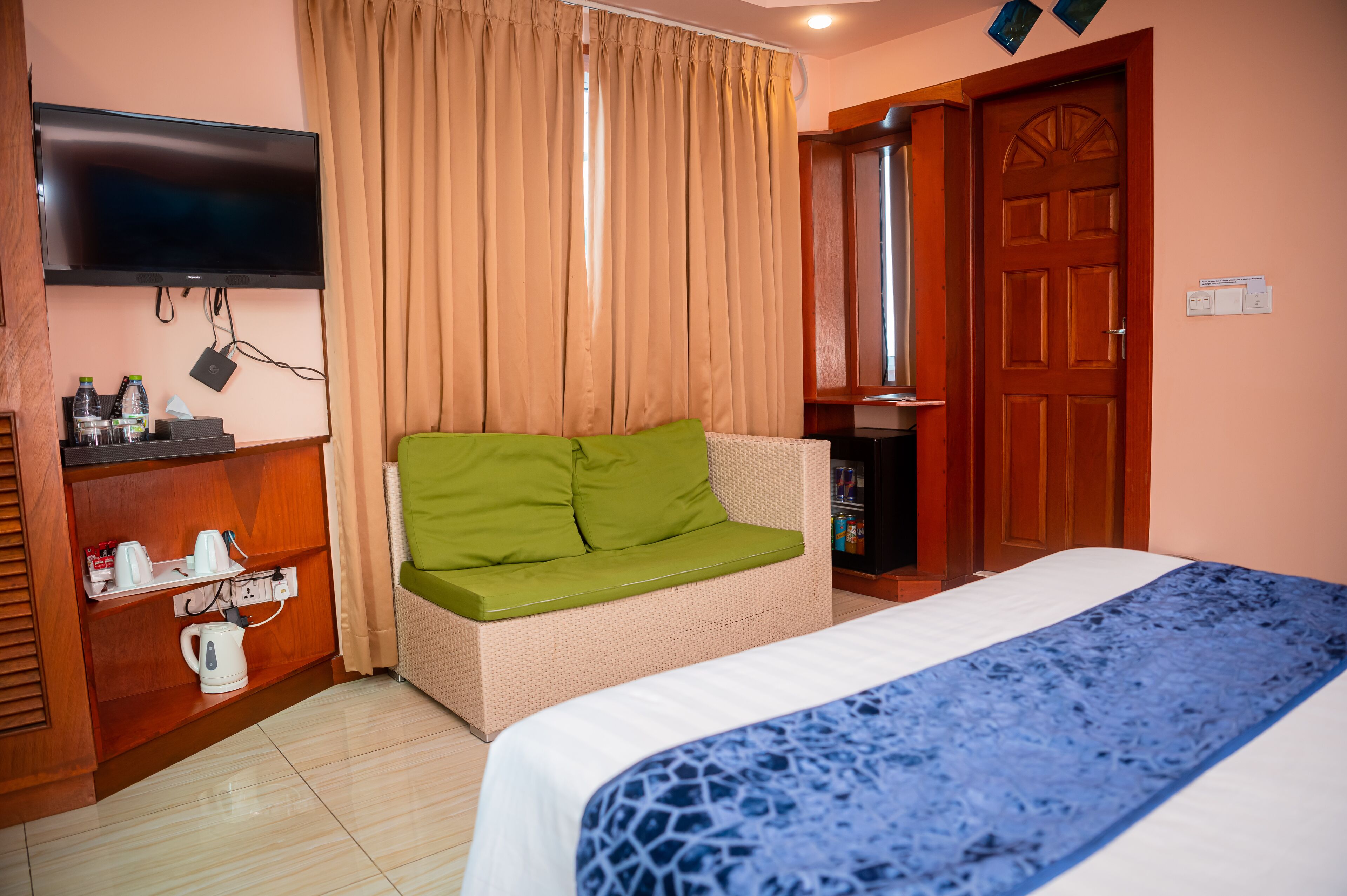 Una habitación de hotel con una cama, un sofá con cojines verdes, un televisor montado en la pared y un mueble de madera con una tetera y otros objetos.