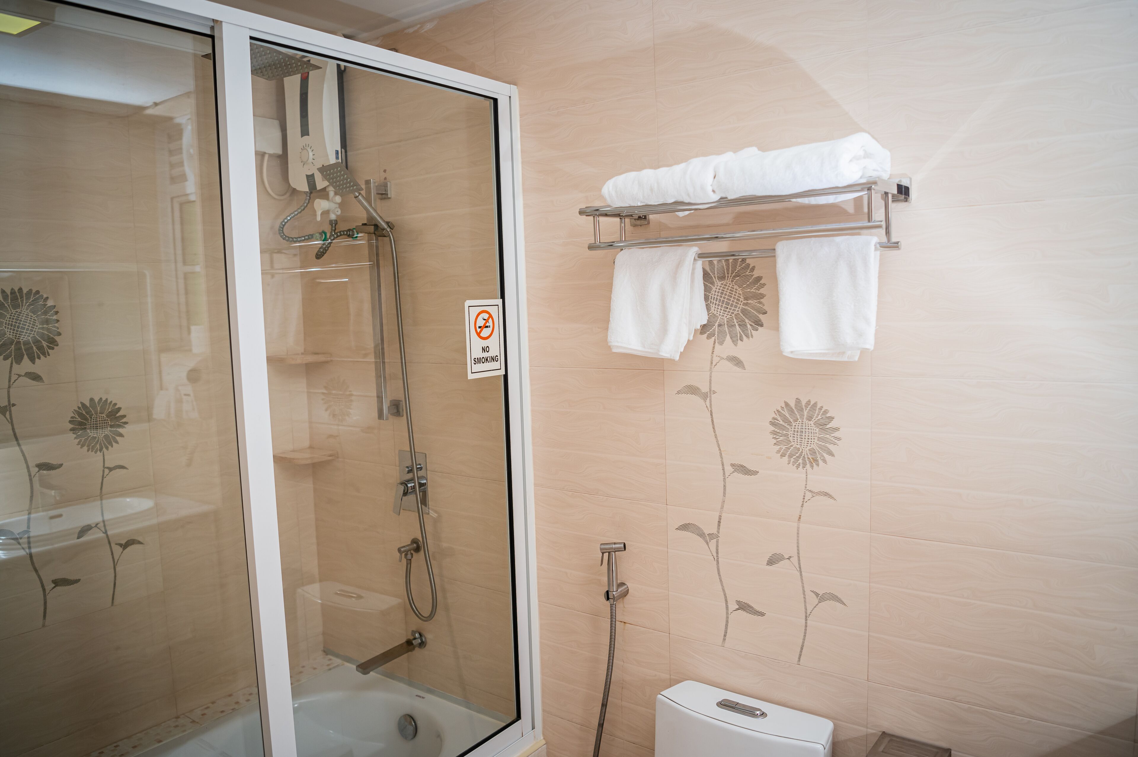 Ein Badezimmer mit einer gläsernen Duschkabine, einem Rauchverbotsschild, einem Handtuchhalter mit Handtüchern und blumigen Wandverzierungen.