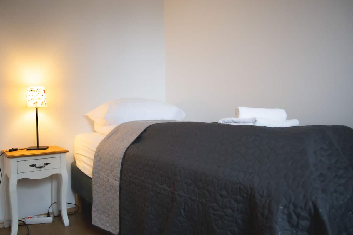 Cama bem arrumada com edredom escuro, travesseiro e manta brancos dobrados ao pé. Uma pequena mesa de cabeceira com abajur e régua de energia é visível.