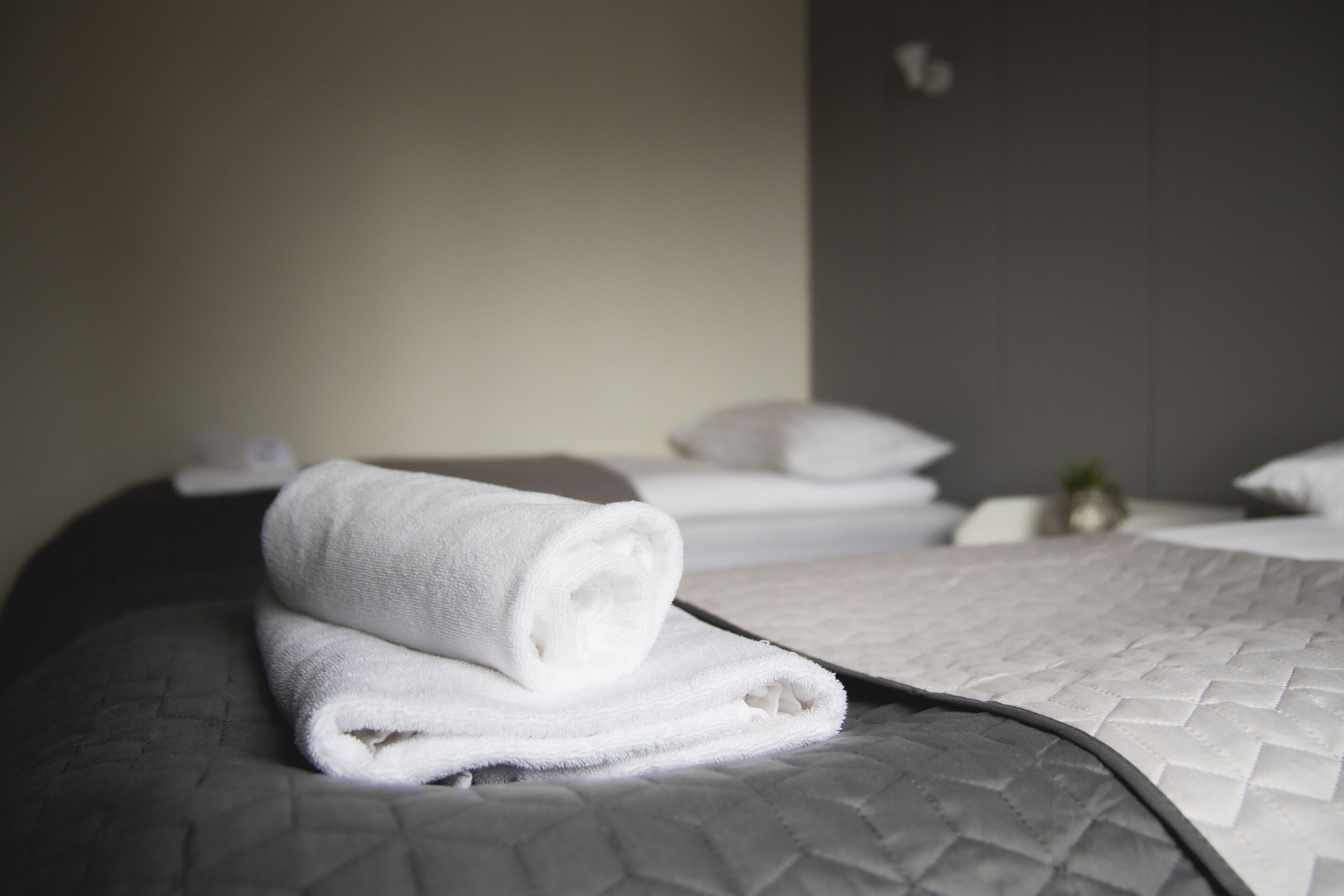 A neatly made bed with a rolled white towel on top, a grey quilted bedspread, and a grey headboard.