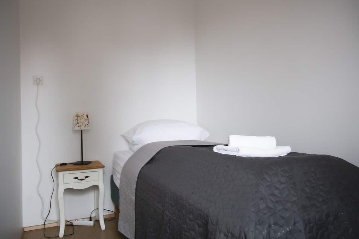 Cama bem arrumada com um edredom escuro, um travesseiro branco e uma toalha branca. Há uma pequena mesa de cabeceira com uma luminária e uma tomada.