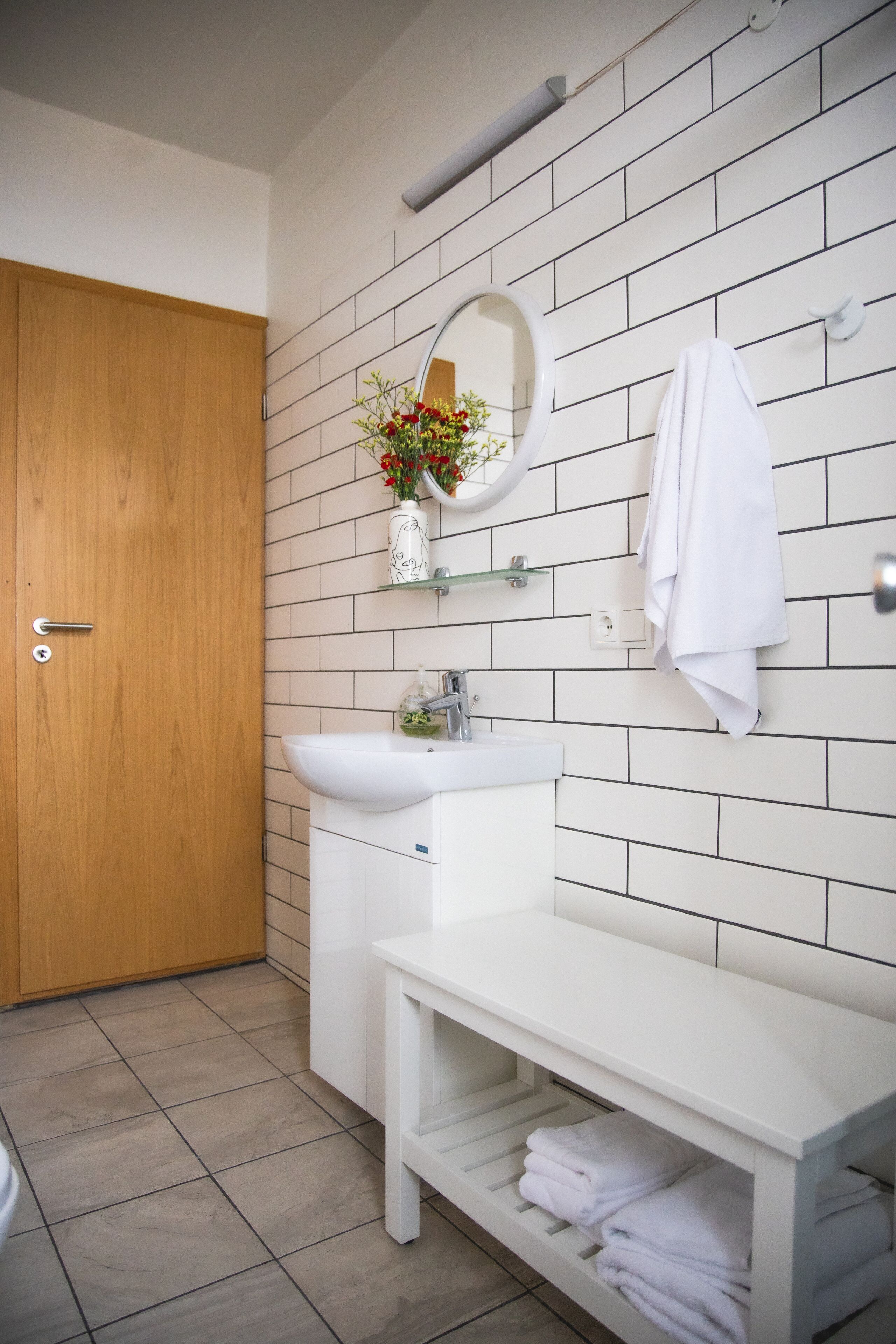 Un bagno con una porta in legno, piastrelle bianche, un lavandino, uno specchio e un portascopevoli.