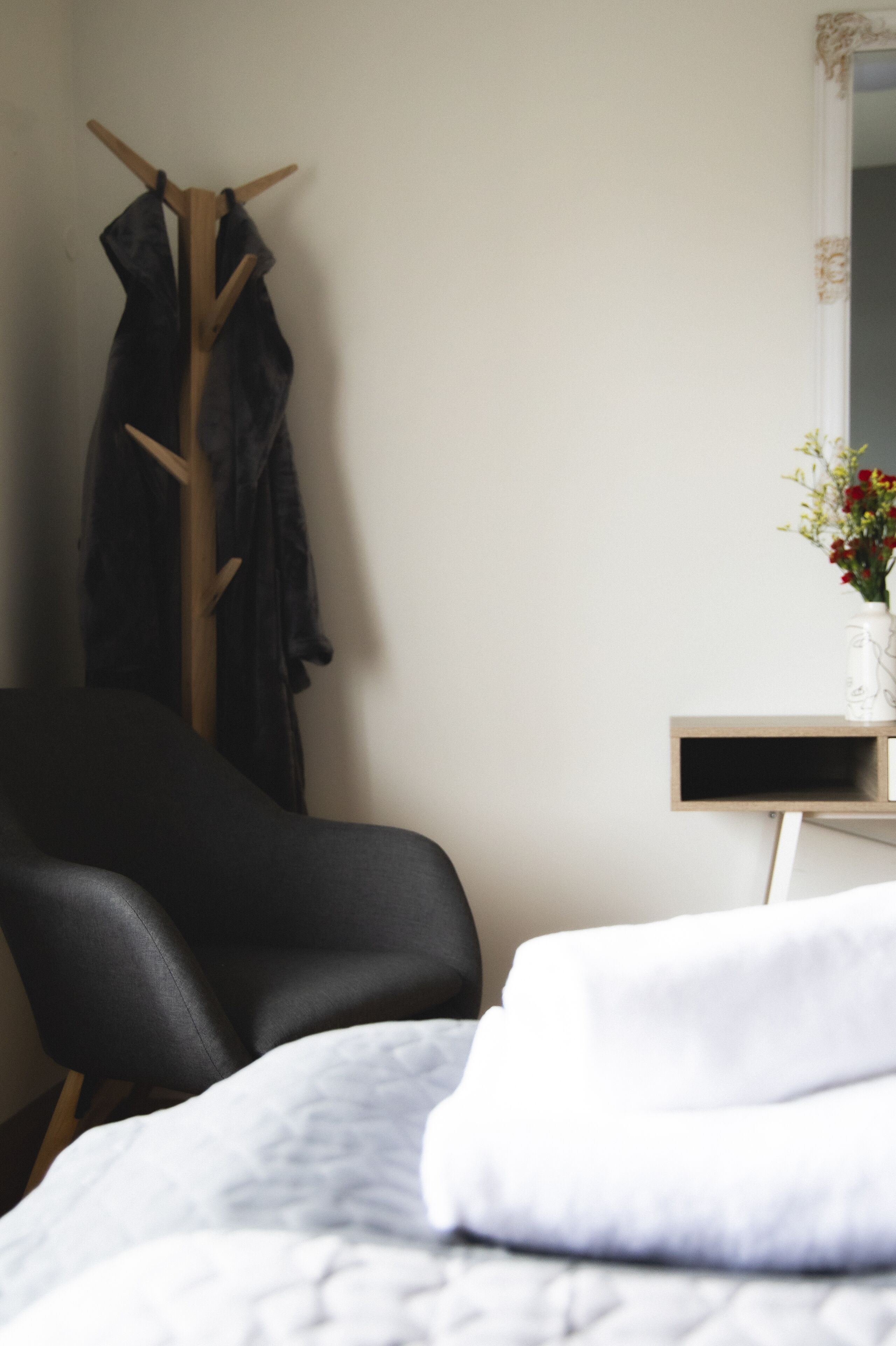 A bedroom with a bed, a chair, a coat rack, and a small table with a vase of flowers.