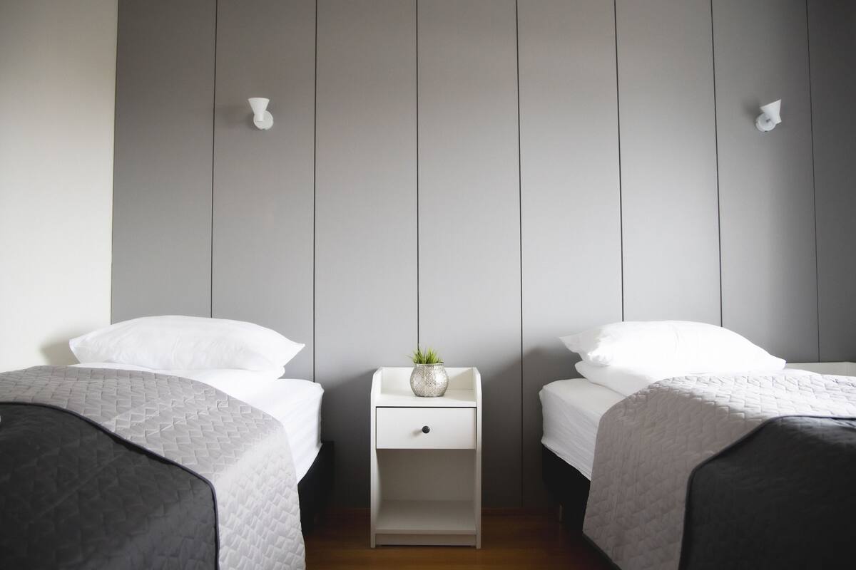 Quarto com duas camas, uma mesinha de cabeceira com gaveta e uma pequena planta.