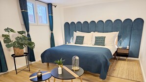 Une chambre à coucher avec un lit, une table de chevet, une commode, une plante en pot, une petite table avec une bougie et une fenêtre avec des rideaux.