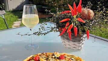 Pizza met diverse groenten op een houten plank, een glas witte wijn en een potplant met rode chilipepers op een buitentafel.