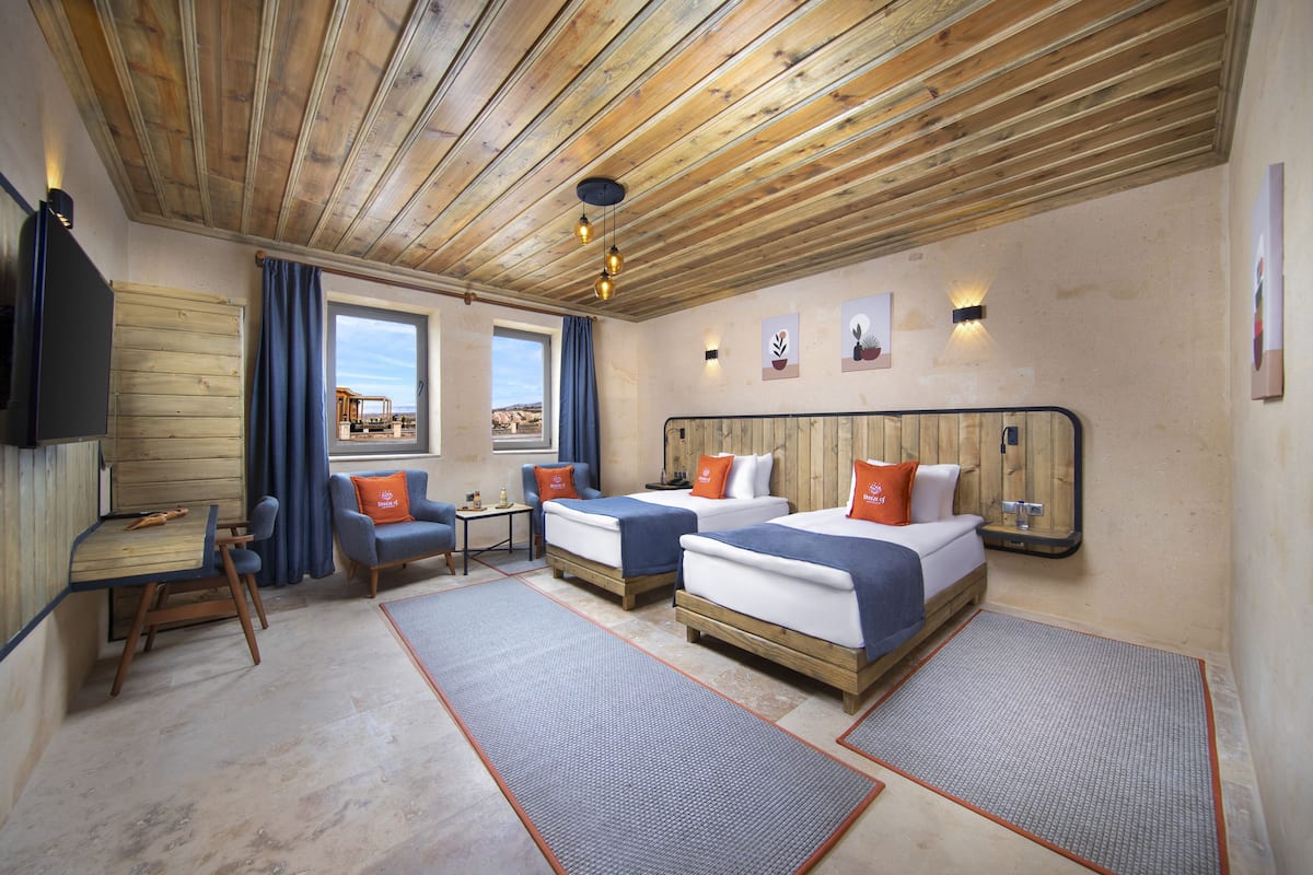 A hotel room with wooden ceiling beams, two beds with blue and orange bedding, a small table with chairs, and a TV mounted on the wall.