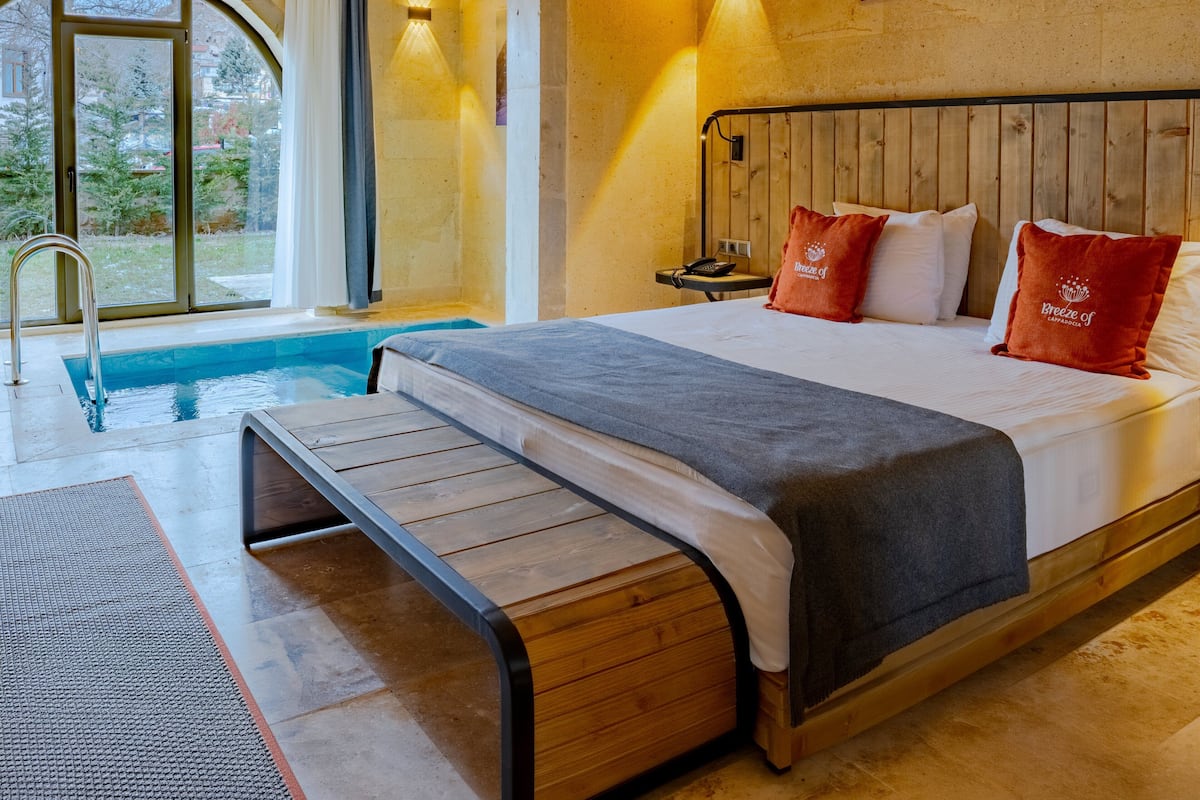 A bedroom with a wooden bed, a small bedside table, a nightstand, a large window, and a view of a swimming pool.