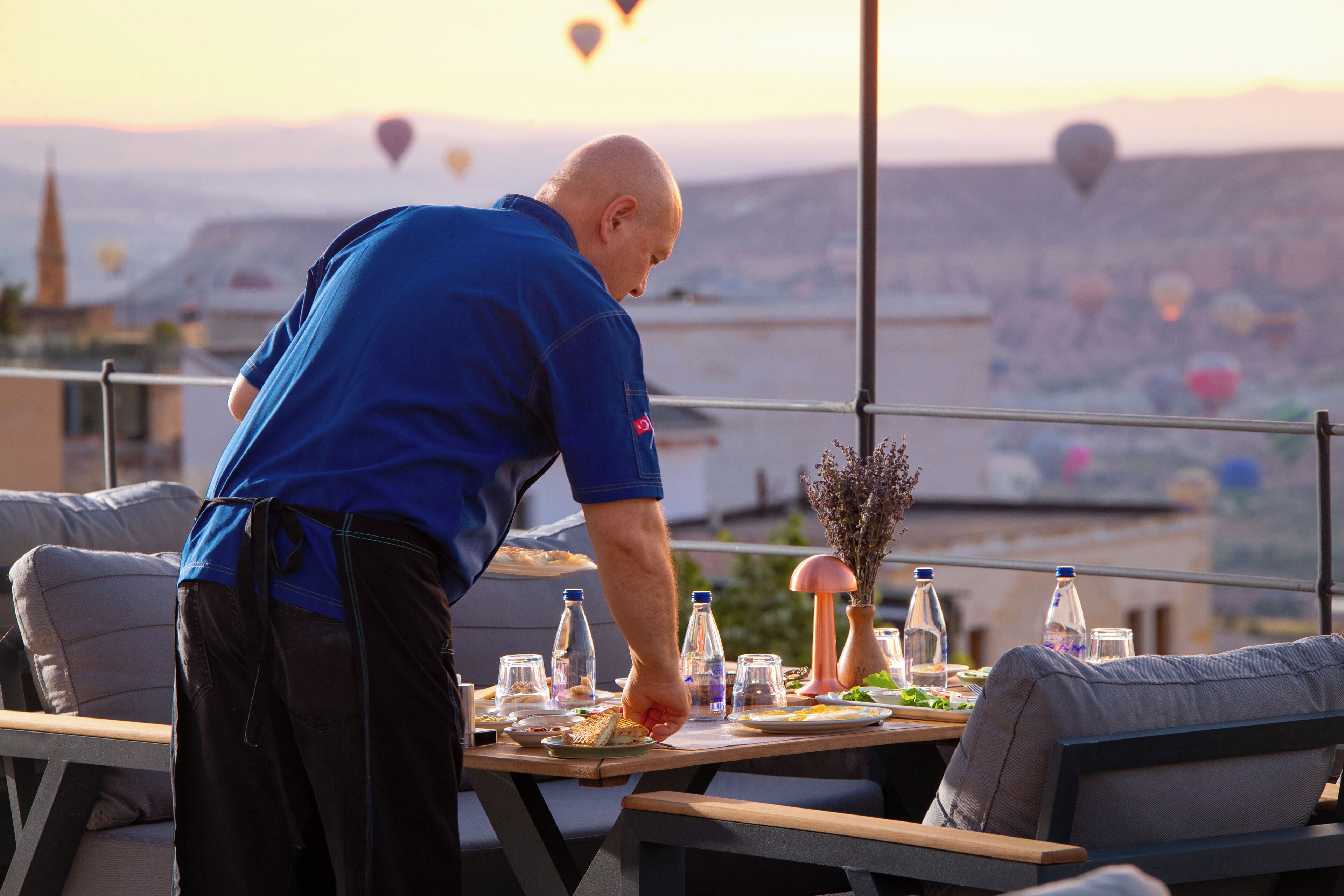 A man serving food at an outdoor table with a city view, hot air balloons, and a sunset in the background.