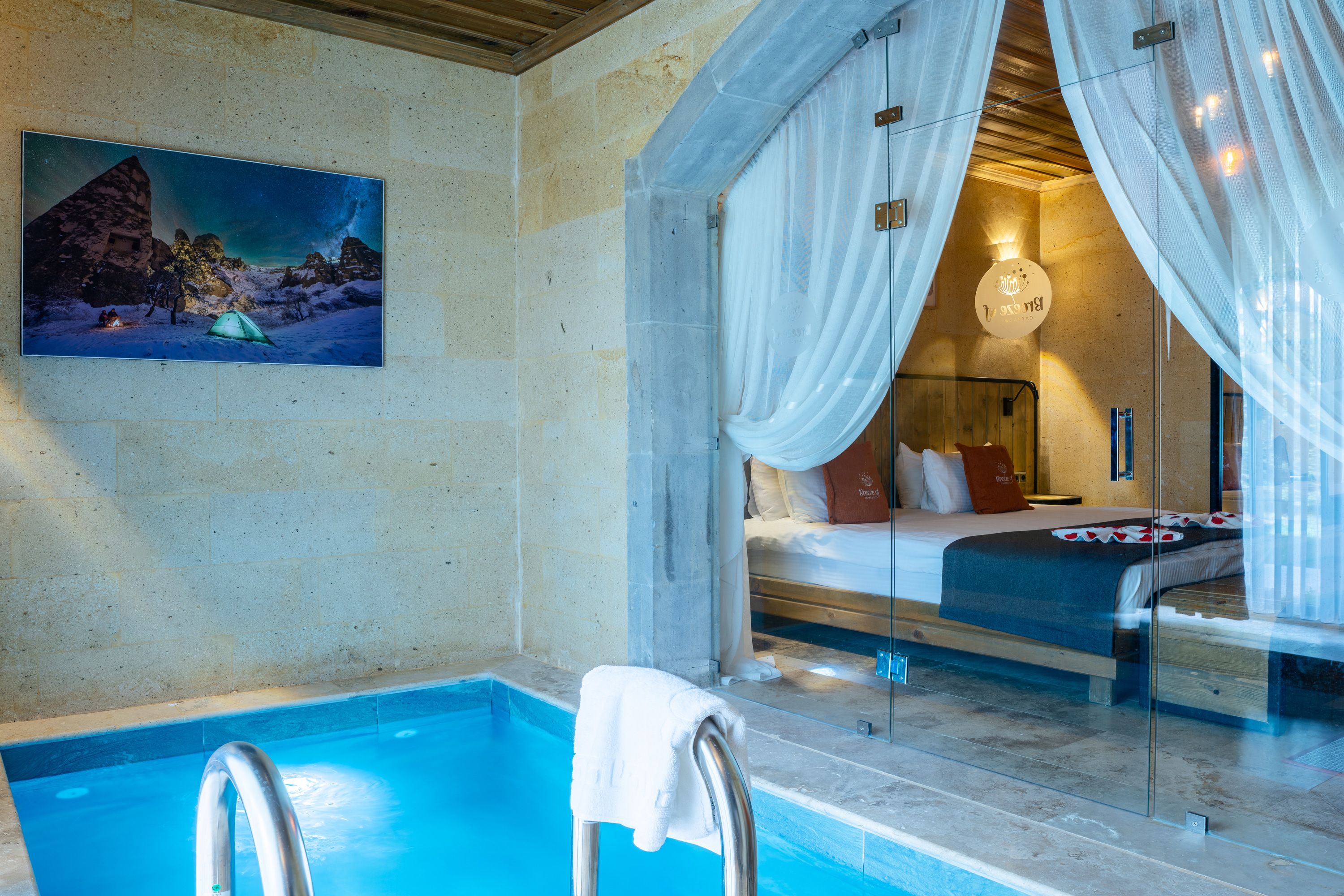 A hotel room with a glass partition, a bed, a bedside table, and a painting on the wall.
