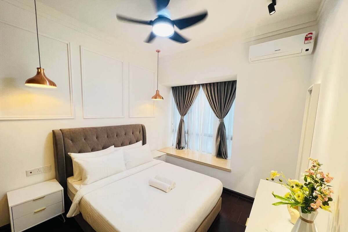 Habitació d'hotel ben ordenada, amb un llit gran, taules de nit, ventilador de sostre, finestra amb cortines i un ram de flors.