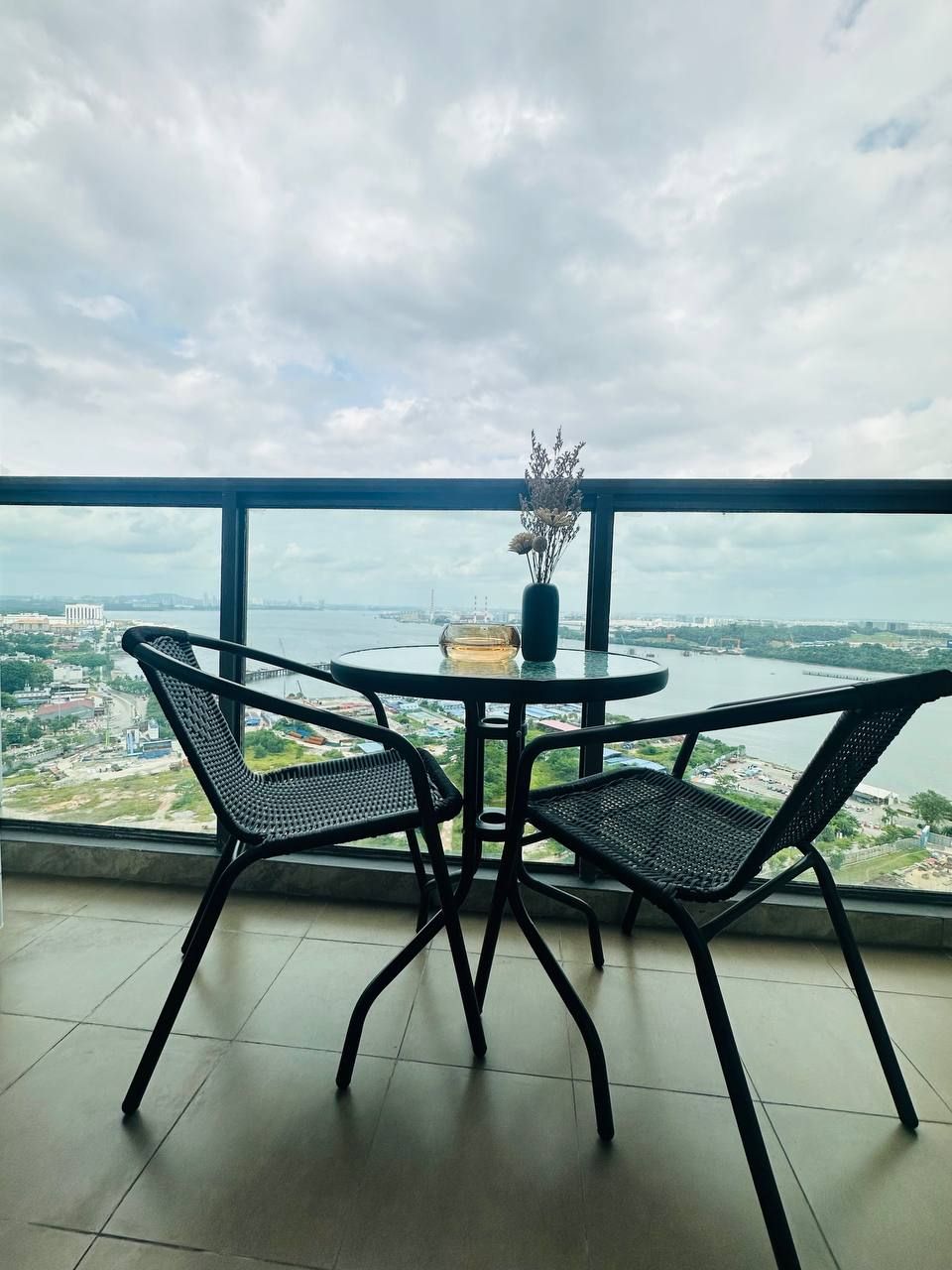 A balcony with two black metal chairs, a small round table, and a vase with dried flowers. The balcony overlooks a body of water and a distant cityscape.