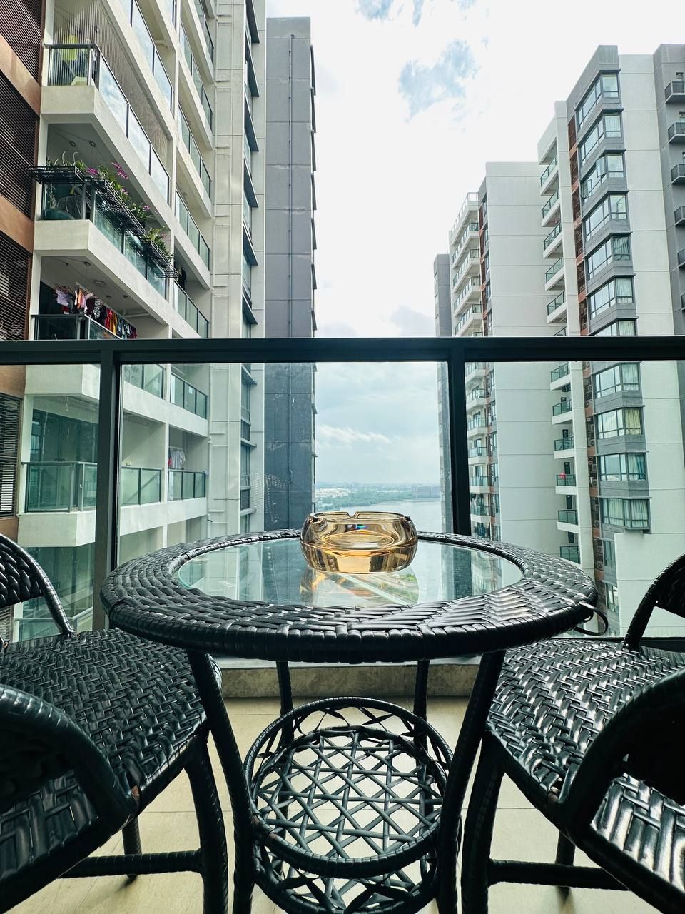 A balcony with a glass table and wicker chairs, overlooking a cityscape.