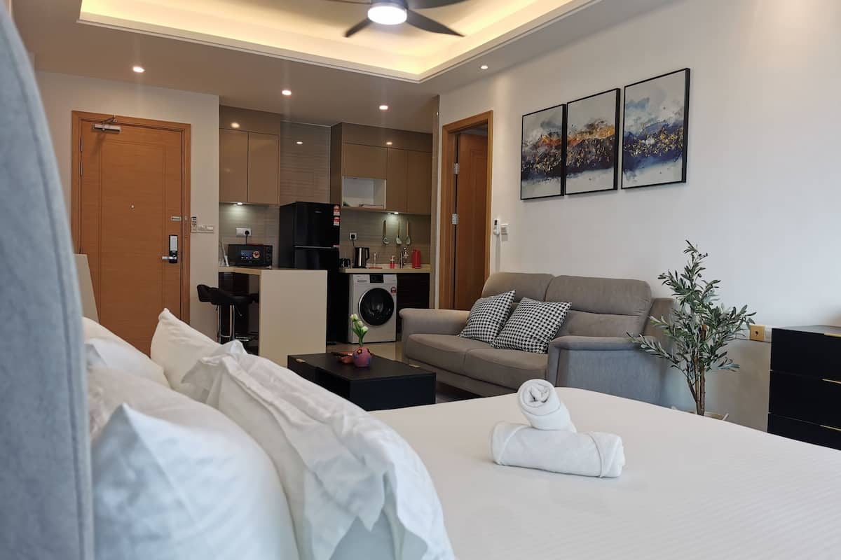 Habitació d'hotel amb llit, sofà, taula petita amb un vas, ventilador de sostre i una cuineta al fons.