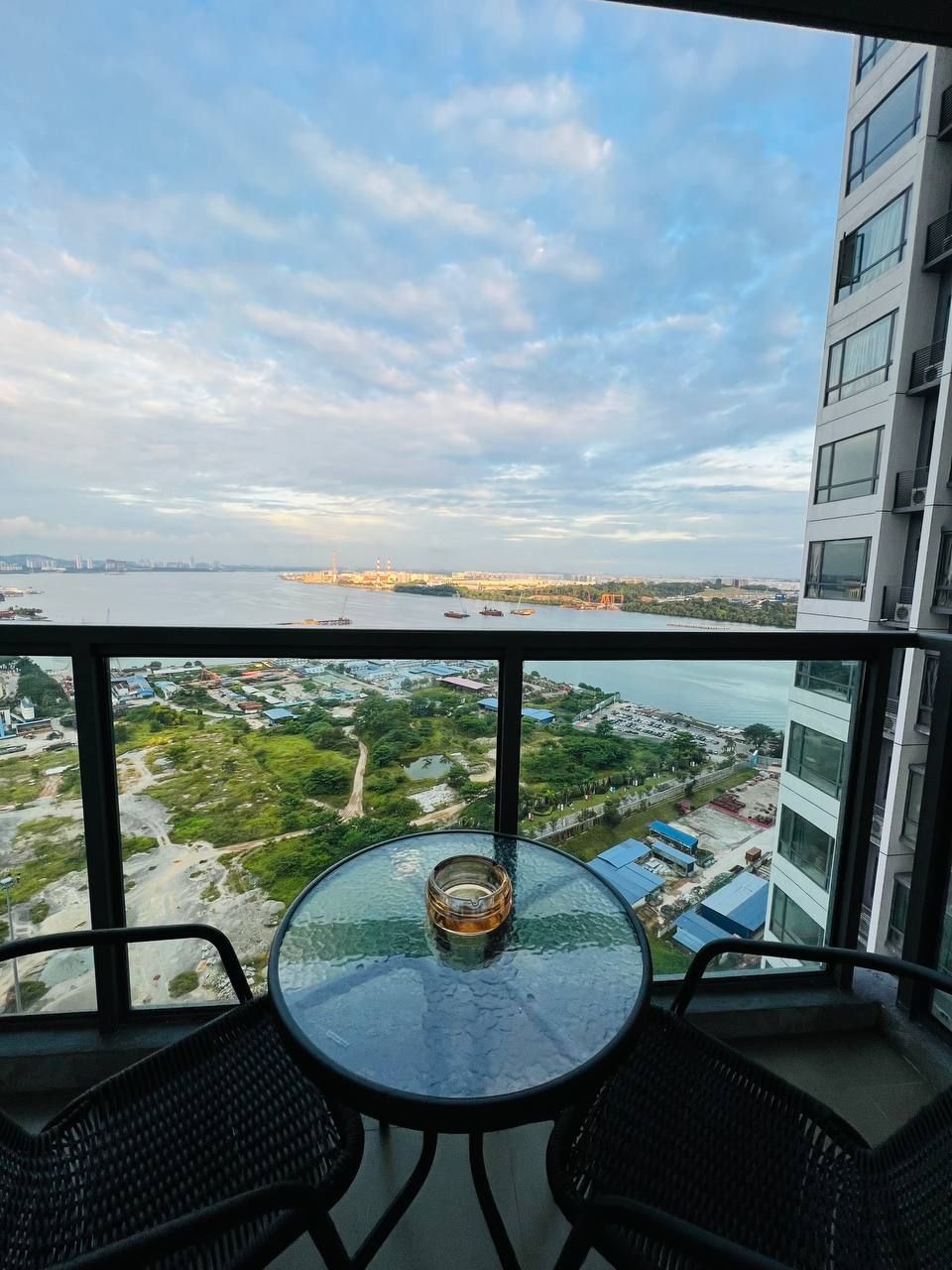 A balcony with a glass table and two chairs, overlooking a river with boats and a distant cityscape.